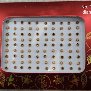 Puede incluir: Una tarjeta blanca con borde rojo y diseños florales dorados. La tarjeta contiene un recipiente de plástico transparente con 100 pequeñas piedras redondas, blancas y con borde dorado. El texto "No. 2,5 mm de diámetro" está impreso en la tarjeta.