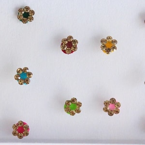 Puede incluir: Surtido de 12 pegatinas bindi de colores en forma de flor con detalles dorados. Las bindis están dispuestas en una cuadrícula sobre un fondo blanco.