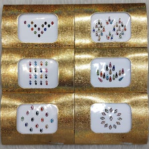 Puede incluir: Seis pequeñas cajas con bordes dorados, cada una contiene una hoja de brillantes adhesivos de colores. Los brillantes están dispuestos en diferentes patrones, incluyendo un corazón, una lágrima y un círculo.