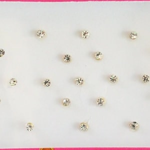 Puede incluir: Un contenedor de plástico blanco con una tapa rosa lleno de pequeños diamantes de imitación transparentes. Los diamantes de imitación están dispuestos en filas sobre un fondo blanco.