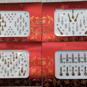 Puede incluir: Cuatro cajas rojas con diseños florales dorados, cada una con joyas faciales. Las joyas son doradas y plateadas, con varias formas y tamaños, incluyendo lágrimas y círculos. Para uso cosmético.