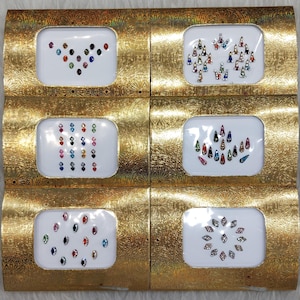 Puede incluir: Seis pequeñas cajas con bordes dorados, cada una contiene una hoja de brillantes adhesivos de colores. Los brillantes están dispuestos en diferentes patrones, incluyendo un corazón, una lágrima y un círculo.