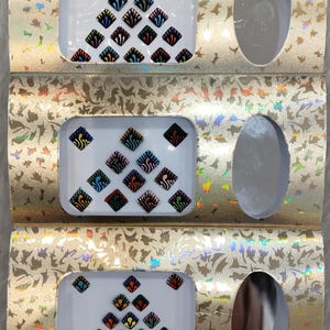 Puede incluir: Tres paquetes de pegatinas bindi decorativas de color en forma de diamante. Cada paquete contiene una variedad de diseños con contornos negros y acentos de colores. Las pegatinas bindi están dispuestas en forma de triángulo.