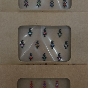 May include: Three rectangular brown cardboard packages containing adhesive face stickers. The top pack features black arrow-like designs with colorful rhinestones. The middle pack shows black paisley shapes with small gems. The bottom pack displays red geometric patterns with crystals.