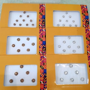 Könnte beinhalten: Sechs kleine Boxen mit gelben Deckeln und weißen Innenräumen. Jede Box enthält einen Satz kleiner, runder, dekorativer Aufkleber mit einem glitzernden Finish. Die Aufkleber sind in einem dreieckigen Muster angeordnet. Drei Boxen enthalten silberne Aufkleber und drei Boxen enthalten goldene Aufkleber.