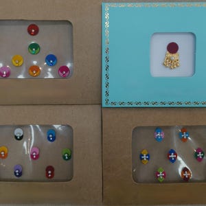 May include: Four rectangular display cards containing adhesive bindi stickers. Three cards are brown with clear windows showing round and oval bindis in various colors like red, green, yellow, blue, and pink. One card is teal with a gold-tone bindi.