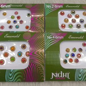 Può includere: Quattro confezioni di Nidhi Bindis, ciascuna con un assortimento diverso di adesivi bindi colorati e dai toni gioiello. Le confezioni sono etichettate con misure da 1 a 8 mm. Il testo "Emerald" è stampato su ogni confezione.