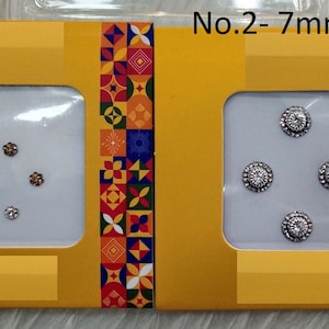 Puede incluir: Dos cajas amarillas con un patrón colorido en la parte delantera. Las cajas contienen dos juegos de pequeños diamantes de imitación redondos de color plateado. El texto en las cajas dice "No. 1 - 3mm" y "No. 2 - 7mm".