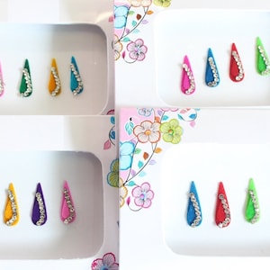 Puede incluir: Un conjunto de 12 pegatinas bindi de colores en forma de lágrima con diamantes de imitación. Los bindis están dispuestos en cuatro filas de tres, cada fila con una combinación de colores diferente.