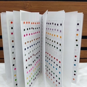 Puede incluir: Un archivador blanco con páginas de pequeños puntos de colores, cada uno de 3,5 mm de diámetro. El archivador está abierto para revelar una variedad de colores, incluyendo rojo, azul, verde, amarillo, naranja, morado y marrón.