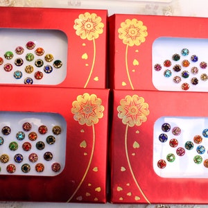 Puede incluir: Cuatro cajas rojas con diseños dorados y ventanas transparentes que muestran brillantes de colores con tonos de piedras preciosas. Los brillantes están dispuestos en forma de diamante.