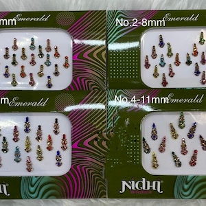 Puede incluir: Cuatro cajas de pegatinas bindi de color esmeralda, cada una con un rango de tamaño diferente: 1-8mm, 2-8mm, 3-10mm y 4-11mm. Las pegatinas están dispuestas en forma de lágrima y vienen en varios colores. La marca "NIDHI" es visible.