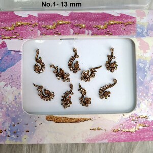 Puede incluir: Un conjunto de ocho pegatinas bindi decorativas de color dorado. Las pegatinas están dispuestas en un marco rectangular blanco sobre un fondo rosa y morado. El texto "No. 1-13 mm" está impreso encima del marco.