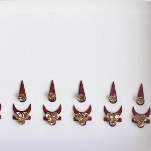 Puede incluir: Seis pegatinas bindi decorativas rojas y doradas con diamantes de imitación. Las pegatinas bindi están dispuestas en una fila sobre un fondo blanco.