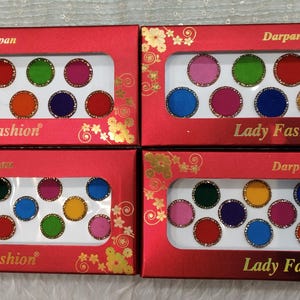 Könnte beinhalten: Vier rote Schachteln mit Lady Fashion Bindi-Punkten in verschiedenen Farben. Jede Schachtel enthält eine Reihe farbiger, runder Bindi-Punkte. Die Schachteln haben goldene Blumendesigns und das Wort "Darpan" oben.