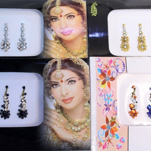 Op de afbeelding: Vier sets van drie bejeweled bindi stickers. Elk set heeft een ander kleurenschema: zilver, goud, zwart en veelkleurig. De bindi stickers zijn gerangschikt op witte kaarten met een vrouwengezicht op de achtergrond.
