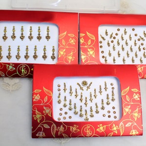 Puede incluir: Tres cajas rojas con diseños florales dorados y bordes blancos. Cada caja contiene una hoja de diamantes de imitación y joyas adhesivas doradas y transparentes. Los diamantes de imitación están dispuestos en varios patrones.