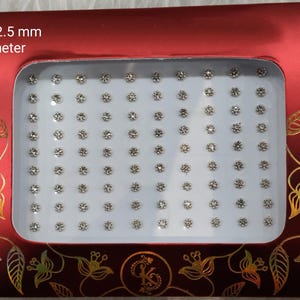 Può includere: Un contenitore di plastica bianco con un coperchio di plastica trasparente pieno di piccoli strass rotondi argentati. Il contenitore ha uno sfondo a motivi rossi e dorati con il testo "No. 1 - 2.5 mm di diametro".