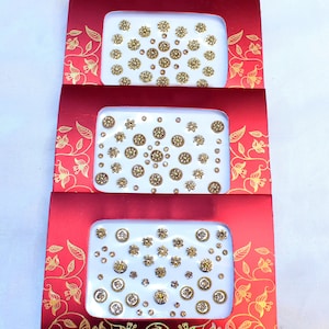 Könnte beinhalten: Drei rote Boxen mit weißen Deckeln, die goldene und klare Strasssteine enthalten. Die Boxen sind mit goldenen Blumendesigns verziert.