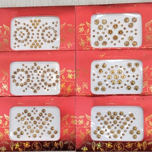 May include: Six packages of face jewels are shown. Each package has a red background with gold floral designs. The face jewels are arranged on a white background in various patterns. The jewels are gold and clear, with some in a flower shape.