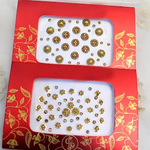 Könnte beinhalten: Zwei rote Schachteln mit goldenen floralen Designs, die jeweils dekorative Verzierungen enthalten. Die obere Schachtel enthält goldene, rote und grüne kreisförmige Verzierungen. Die untere Schachtel enthält goldene und braune kreisförmige Verzierungen.