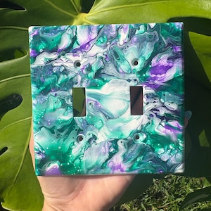 Puede incluir: Una cubierta de interruptor doble cuadrada con un diseño de arte fluido. La cubierta presenta un patrón arremolinado de pintura turquesa, morada y blanca. Tiene dos recortes rectangulares para los interruptores y dos orificios para tornillos.