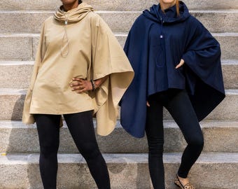 May include: Two hooded ponchos, one in beige and one in navy blue, each featuring a drawstring neckline with a toggle closure. The ponchos have a loose, draped silhouette with wide sleeves and are worn over black leggings with flat shoes.