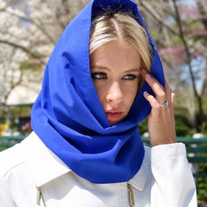 May include: A vibrant royal blue hood made of smooth fabric draped over the head, featuring a structured cowl neck design that frames the face. The hood is worn over a crisp white jacket with gold-toned metal zippers visible on the front panels.