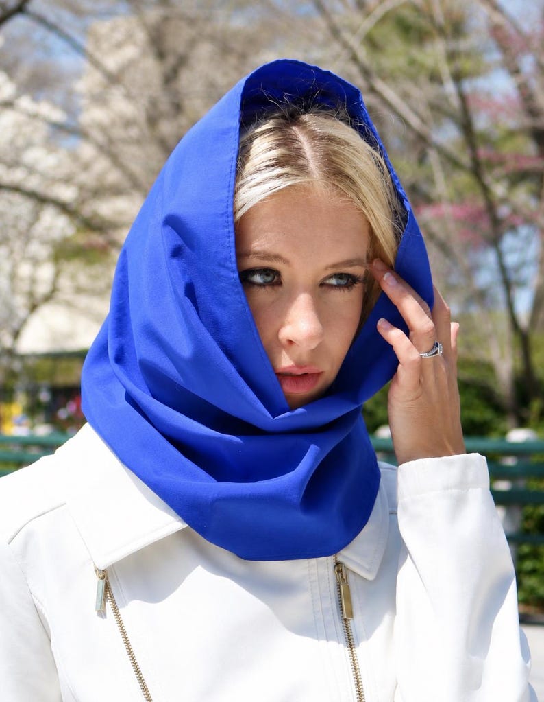 May include: A vibrant royal blue hood made of smooth fabric draped over the head, featuring a structured cowl neck design that frames the face. The hood is worn over a crisp white jacket with gold-toned metal zippers visible on the front panels.