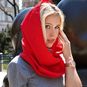 May include: A bright red hooded scarf worn over a gray short-sleeved t-shirt. The scarf features a solid, vibrant red fabric that drapes around the head and neck, providing a bold contrast against the neutral gray shirt worn underneath.