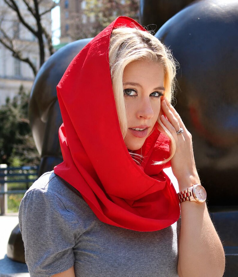 May include: A bright red hooded scarf worn over a gray short-sleeved t-shirt. The scarf features a solid, vibrant red fabric that drapes around the head and neck, providing a bold contrast against the neutral gray shirt worn underneath.