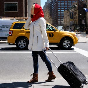 May include: A person wearing a bright red hooded scarf, a cream-colored zip-up jacket, dark blue jeans, and brown lace-up boots. The person is pulling a black rolling suitcase with a retractable handle while walking along a city street.