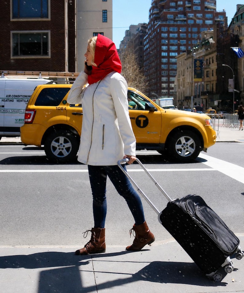 May include: A person wearing a bright red hooded scarf, a cream-colored zip-up jacket, dark blue jeans, and brown lace-up boots. The person is pulling a black rolling suitcase with a retractable handle while walking along a city street.