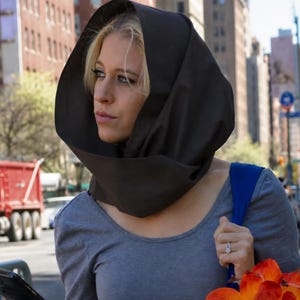 May include: A dark brown fabric hood worn over the head, featuring a structured, rounded shape that frames the face. The hood is made of a solid, opaque material and sits loosely around the neck and shoulders, providing full head coverage.