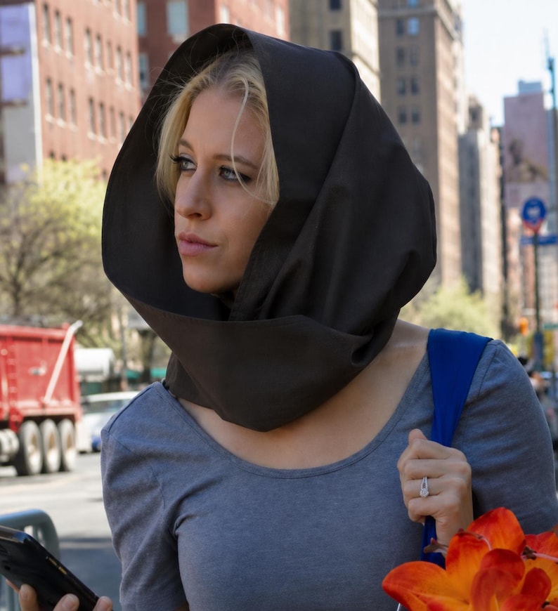 May include: A dark brown fabric hood worn over the head, featuring a structured, rounded shape that frames the face. The hood is made of a solid, opaque material and sits loosely around the neck and shoulders, providing full head coverage.
