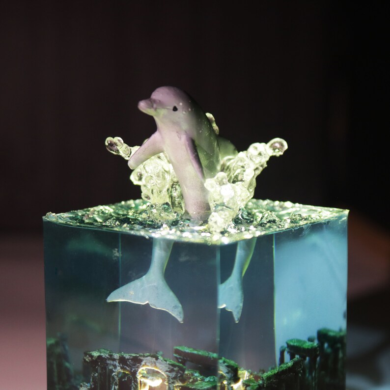 May include: A clear resin cube with a small gray dolphin figurine inside. The dolphin is leaping out of the water, which is represented by the clear resin. The cube is sitting on a dark surface.