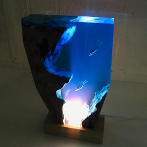 K&ouml;nnte beinhalten: Eine einzigartige Tischlampe mit blauem Harzdesign, das Taucher in einem dunklen Holzrahmen zeigt. Die Lampe strahlt ein sanftes Leuchten aus und erzeugt eine Unterwasserszene. Die Lampe ist auf einem Holzsockel montiert. Abmessungen nicht sichtbar.
