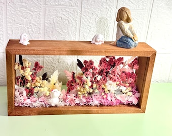 May include: A wooden shadow box filled with dried flowers in shades of pink, white, and yellow. The box is adorned with small white bunny figurines and a figurine of a girl. The box is set on a light green surface.