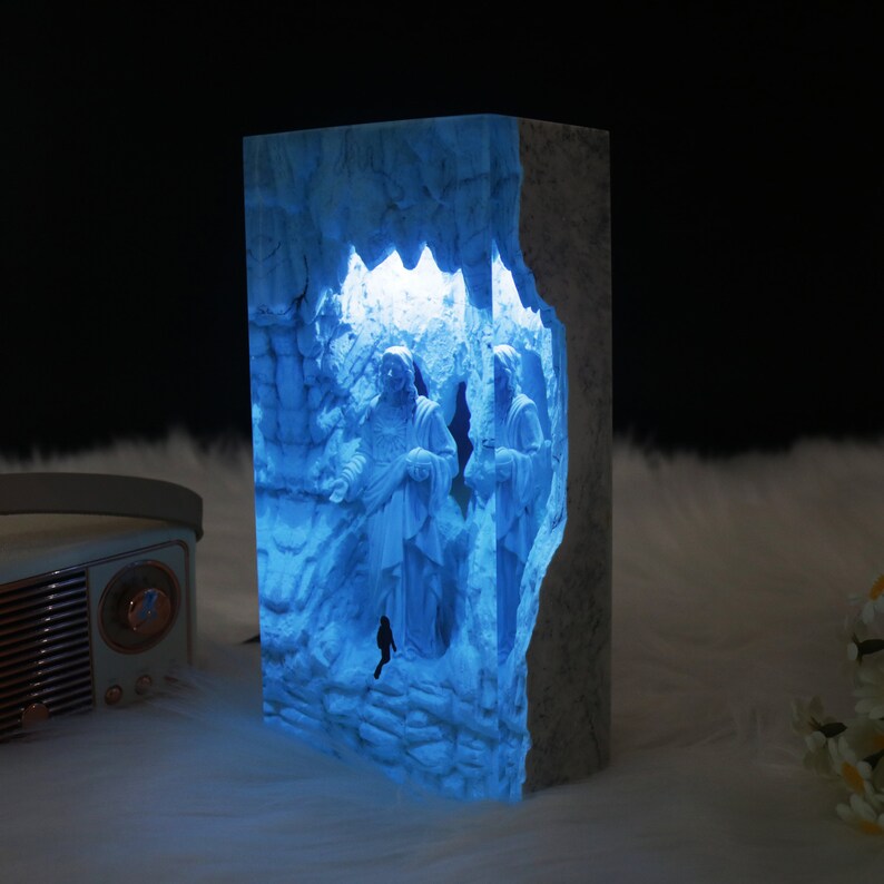 May include: A blue resin lamp with a 3D design of three figures standing in a cave. The lamp is lit from within, casting a blue glow.