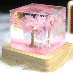 Puede incluir: Un cubo de vidrio transparente con un árbol de cerezo en flor rosa y dos figuritas de ciervos blancos en su interior. El cubo se coloca sobre una base de madera.