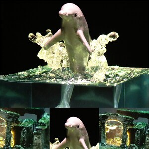 May include: A small, gray dolphin figurine leaping out of a clear, blue resin base. The base is textured to resemble water with bubbles and waves. The dolphin is positioned in a jumping pose with its tail extended behind it.