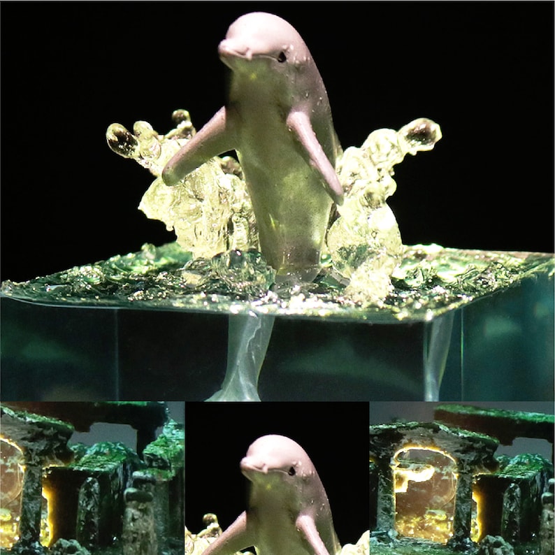 May include: A small, gray dolphin figurine leaping out of a clear, blue resin base. The base is textured to resemble water with bubbles and waves. The dolphin is positioned in a jumping pose with its tail extended behind it.
