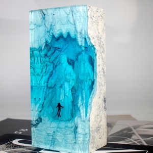 May include: A blue resin sculpture with a white wood frame. The resin is translucent and has a swirling pattern. There are two small figures inside the resin.