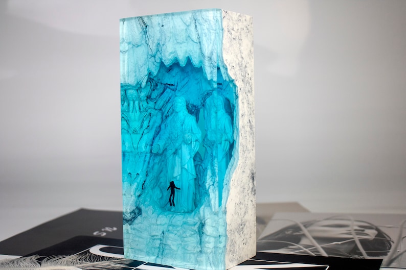 May include: A blue resin sculpture with a white wood frame. The resin is translucent and has a swirling pattern. There are two small figures inside the resin.