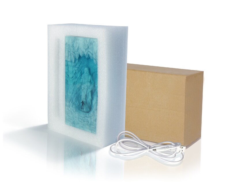 May include: A blue and white 3D printed light-up sculpture of a person standing in a frozen landscape. The sculpture is packaged in a white foam box with a brown cardboard box and a white USB cable.