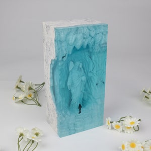 May include: A rectangular block of clear blue resin with white swirls and a white border. Inside the resin is a 3D image of an angel with wings and a person walking towards the angel.