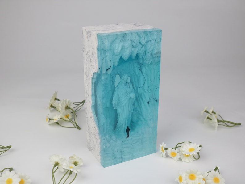 May include: A rectangular block of clear blue resin with white swirls and a white border. Inside the resin is a 3D image of an angel with wings and a person walking towards the angel.