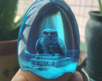 Könnte beinhalten: Eine blaue, eiförmige Kunstharzskulptur mit einer detaillierten Eule, die auf einem Bücherstapel sitzt. Die Eule und die Bücher sind dunkelblau und in einem durchscheinenden, hellblauen Kunstharz eingeschlossen. Der Sockel ist hellbraun.