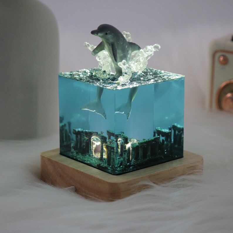 May include: A clear resin cube with a dolphin figurine jumping out of the water. The cube is on a wooden base and has a blue and green tint. The dolphin is gray and has a white belly.