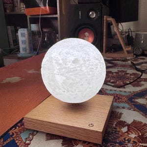 May include: A levitating moon lamp with a textured surface, hovering above a wooden base. The base has a power button. The lamp emits a soft, white glow, creating a celestial ambiance. The lamp is a decorative item.
